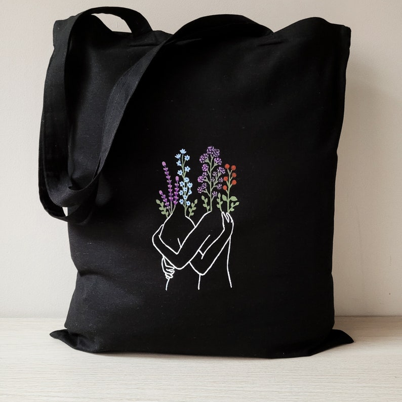 May include: Black tote bag with a white outline of two figures embracing, their heads replaced with colorful flowers.
