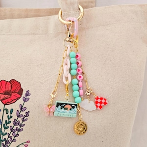 May include: A gold-toned keychain featuring a light pink plastic link, a strand of mint green beads, a strand of pink heart beads, and a light blue cassette tape charm with a sunset graphic. Includes a pink butterfly, a white cloud, and a red checkered heart.