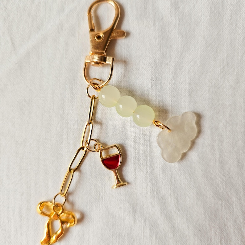 Puede incluir: Un llavero dorado con un dije de copa de vino, un dije de nube y un dije de lazo dorado. El llavero tambi&eacute;n tiene una serie de cuentas de color verde claro.