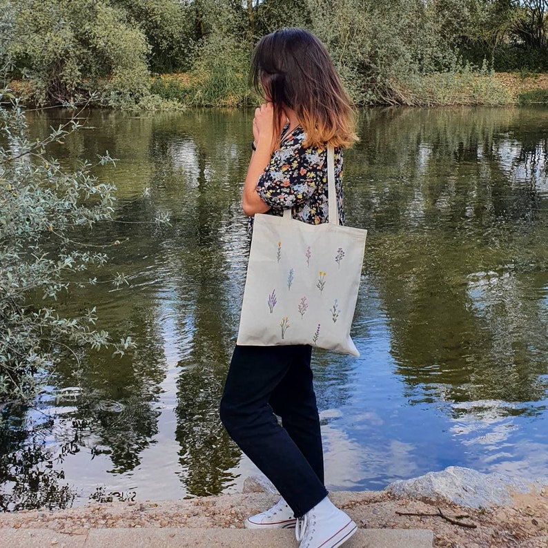 May include: A white tote bag with a floral design. The bag has a long strap and is being held by a person standing near a body of water.