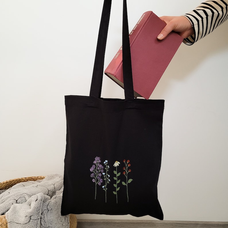 K&ouml;nnte beinhalten: Eine schwarze Tragetasche mit einem Design aus vier verschiedenen Blumen in Lila, Blau, Wei&szlig; und Rot. Die Tasche hat lange schwarze Henkel und ist aus Canvas gefertigt.