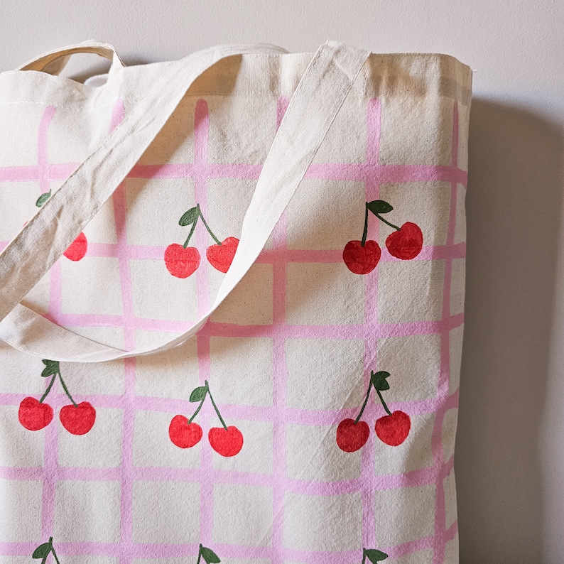 May include: A white canvas tote bag with pink and white checkered pattern and a repeated design of red cherries with green stems.