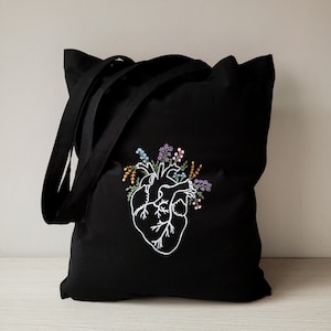 K&ouml;nnte beinhalten: Schwarze Stofftasche aus Canvas mit einem wei&szlig;en gestickten Design eines menschlichen Herzens, aus dem Blumen wachsen.