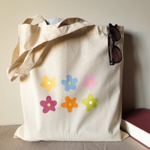 Peut inclure: Un sac fourre-tout en toile beige avec six motifs de fleurs color&eacute;s. Les fleurs sont jaunes, bleues, roses, rouges, oranges et vertes. Une paire de lunettes de soleil noires est accroch&eacute;e au sommet du sac.