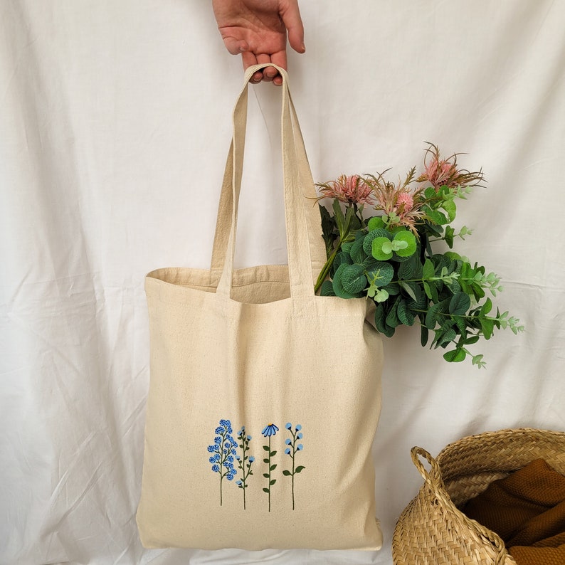Peut inclure: Un sac fourre-tout beige avec un motif floral brod&eacute; sur le devant. Le motif repr&eacute;sente quatre fleurs bleues avec des tiges et des feuilles vertes.
