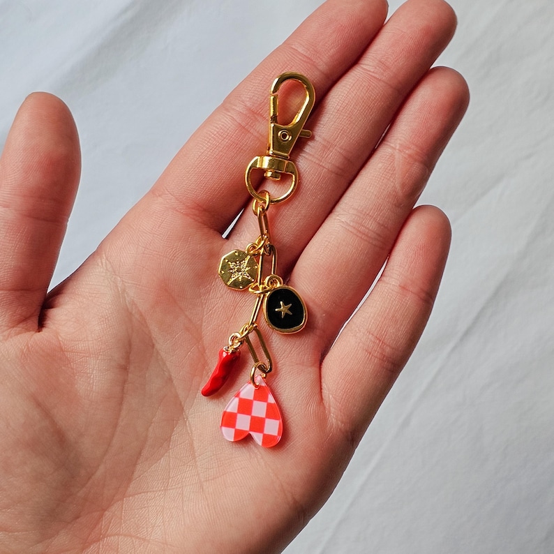 May include: Gold-colored keychain with multiple charms. Charms include a gold-colored starburst, a black charm with a star, a red chili pepper, and a red and white checkered heart. The keychain is held in a hand.