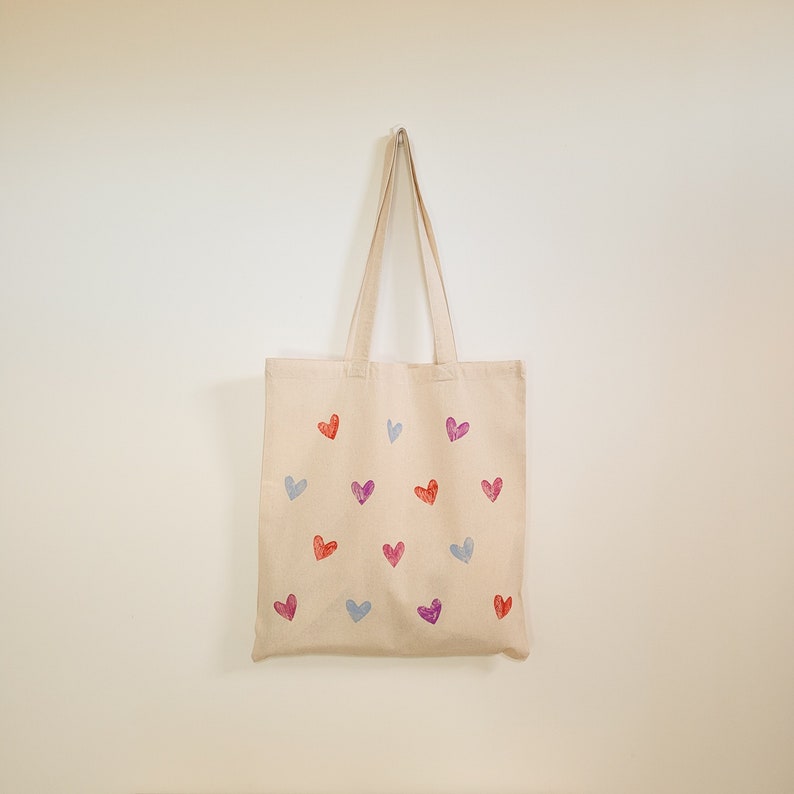 May include: A beige tote bag with a design of colorful hearts printed on it. The bag has two handles and is hanging on a white wall.