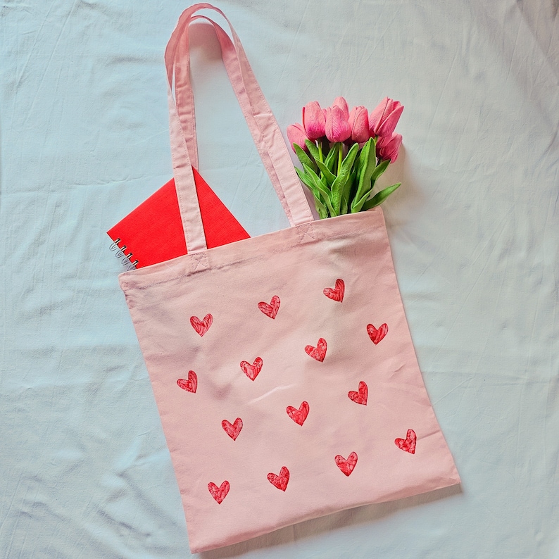 May include: A pink tote bag with a red notebook and pink flowers. The bag has a design of white hearts on it.