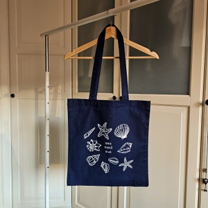 May include: A navy blue tote bag with white illustrations of seashells, starfish, and the text "Sea Sand Sun".