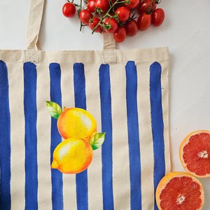 May include: A white canvas tote bag with blue stripes and a painted design of two yellow lemons.