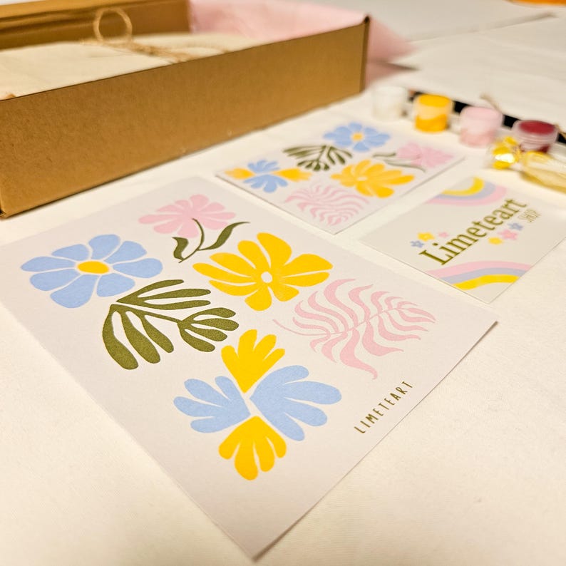 May include: A collection of art prints featuring colorful floral designs in blue, yellow, pink, and green. The prints are arranged on a white surface, with a cardboard box and paint supplies in the background. The text "LIMETEART" is visible on one of the prints.