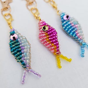 May include: Three beaded fish keychains in shades of blue, pink, and purple. Each fish has a gold-colored clasp and a sequin eye. The fish tails are yellow, and the bodies are made of small, colorful beads.