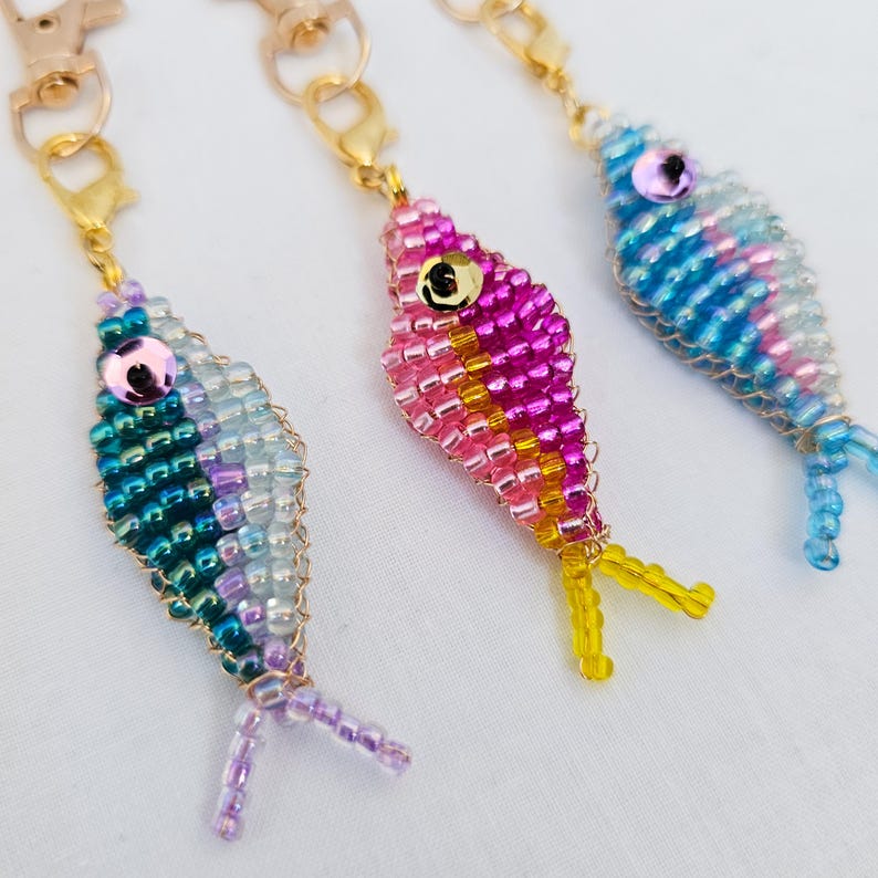May include: Three beaded fish keychains in shades of blue, pink, and purple. Each fish has a gold-colored clasp and a sequin eye. The fish tails are yellow, and the bodies are made of small, colorful beads.