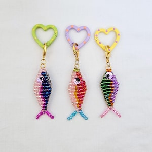 May include: Three beaded fish keychains with heart-shaped clasps. Each fish is a different color, with pink, blue, and purple beads. The clasps are green, purple, and yellow, each with a different color of polka dots. The keychains have gold-colored hardware.