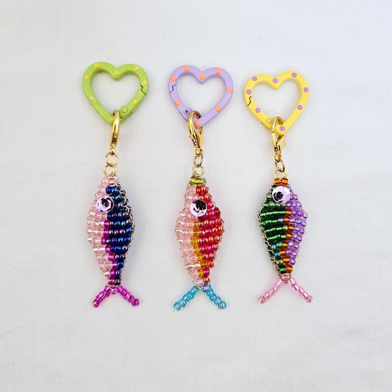 May include: Three beaded fish keychains with heart-shaped clasps. Each fish is a different color, with pink, blue, and purple beads. The clasps are green, purple, and yellow, each with a different color of polka dots. The keychains have gold-colored hardware.