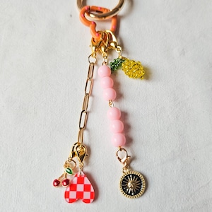 May include: A gold keychain with a pink beaded chain and various charms, including a red and white checkered heart, a lemon, and a sun charm.