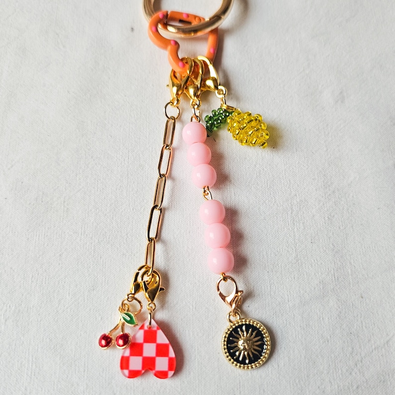 May include: A gold keychain with a pink beaded chain and various charms, including a red and white checkered heart, a lemon, and a sun charm.