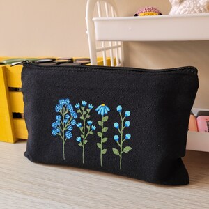 May include: Black canvas zippered pouch with embroidered blue and green flowers. The flowers are stylized and have a simple design.