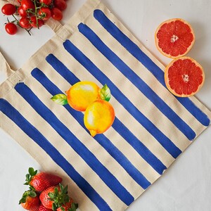 May include: A white canvas tote bag with blue stripes and a painted design of two yellow lemons. The bag has a long handle.