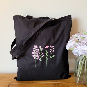 May include: Black canvas tote bag with embroidered pink and green flowers. The bag has a long strap and is ideal for carrying everyday essentials.