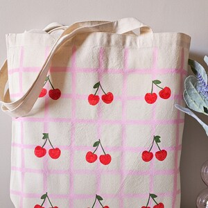 May include: A pink and white canvas tote bag with a repeating pattern of cherries. The cherries are painted in red and green.
