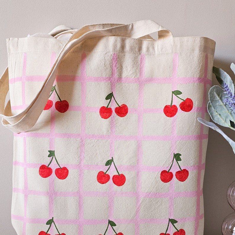 May include: A pink and white canvas tote bag with a repeating pattern of cherries. The cherries are painted in red and green.