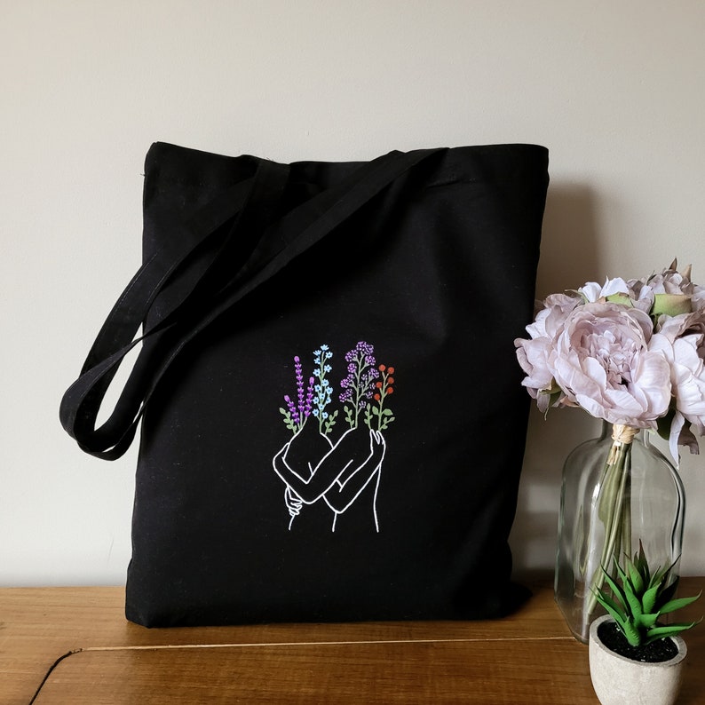 May include: Black tote bag with a white embroidered design of two figures embracing, each with flowers growing from their heads. The flowers are purple, blue, and orange.