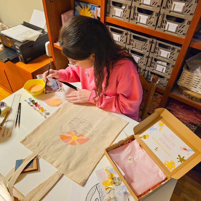 May include: A person painting a design on a beige tote bag with a watercolor design of a sun and mountains. A box with a pink shirt and art supplies is open on the table. The person is wearing a pink sweatshirt.