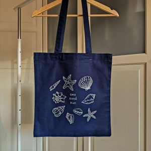 May include: A dark blue tote bag featuring white illustrations of seashells, starfish, and the words "sea sand sun".
