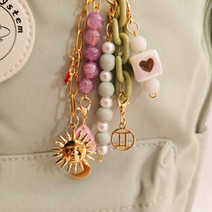 May include: A collection of gold-toned charms and beads, including a sun and moon pendant, pink teardrop, and a Gemini symbol. The charms are attached to a light green chain and a chain of purple and white beads. The background is a light green fabric.