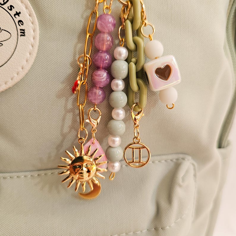 May include: A collection of gold-toned charms and beads, including a sun and moon pendant, pink teardrop, and a Gemini symbol. The charms are attached to a light green chain and a chain of purple and white beads. The background is a light green fabric.