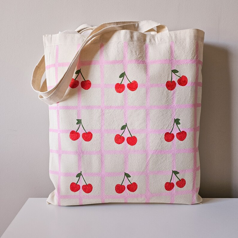 May include: A white canvas tote bag with pink checkered pattern and red cherries printed on it.
