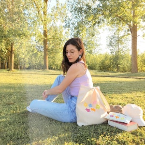 Peut inclure: Une femme portant un d&eacute;bardeur violet clair et un jean bleu est assise sur l'herbe avec un sac cabas blanc brod&eacute; de fleurs color&eacute;es. Le sac est &agrave; c&ocirc;t&eacute; d'un livre ray&eacute; brun et blanc et d'un sac cabas en toile blanc.