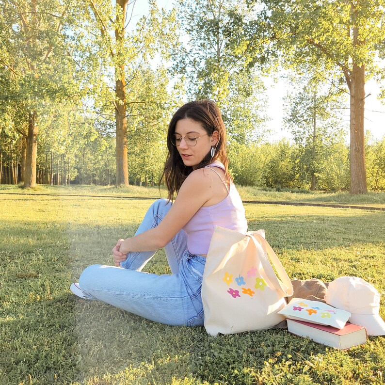 Peut inclure: Une femme portant un d&eacute;bardeur violet clair et un jean bleu est assise sur l'herbe avec un sac cabas blanc brod&eacute; de fleurs color&eacute;es. Le sac est &agrave; c&ocirc;t&eacute; d'un livre ray&eacute; brun et blanc et d'un sac cabas en toile blanc.