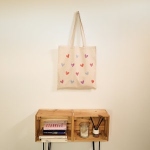 May include: A beige tote bag with a pattern of pink, purple, and blue hearts. The bag is hanging on a white wall above a wooden shelf with books and decorative items.