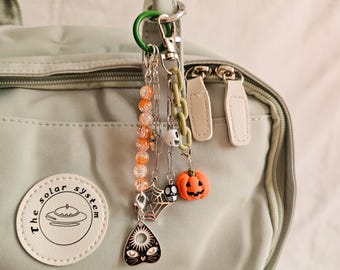 May include: A collection of Halloween-themed charms hanging from a green clasp. Charms include a jack-o'-lantern, a skull, a spider web, and a black cat pendant. The charms are attached to a light green bag with a patch that says "the solar system".