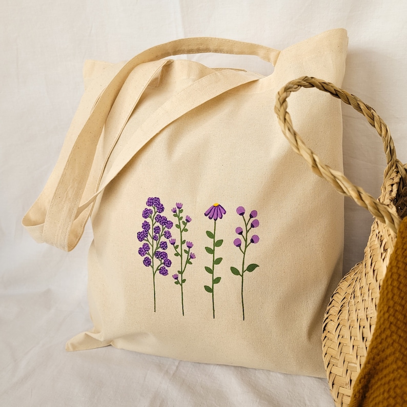 May include: A beige canvas tote bag with purple floral embroidery. The flowers are stylized and include a daisy, a cluster of small flowers, and two sprigs of berries.