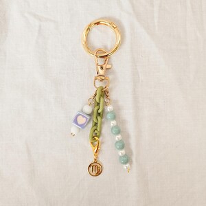 May include: Gold-tone keychain with a lobster clasp and a large ring. The keychain features a light green chain, a charm with a Virgo symbol, and strands of beads in white, light green, and a square bead with a heart.