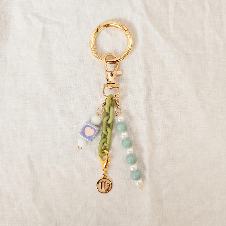 May include: Gold-tone keychain with a lobster clasp and a large ring. The keychain features a light green chain, a charm with a Virgo symbol, and strands of beads in white, light green, and a square bead with a heart.