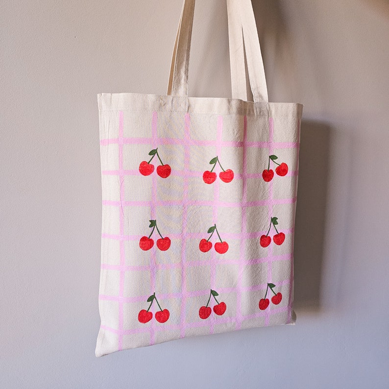 May include: A white canvas tote bag with pink checkered pattern and red cherry illustrations.