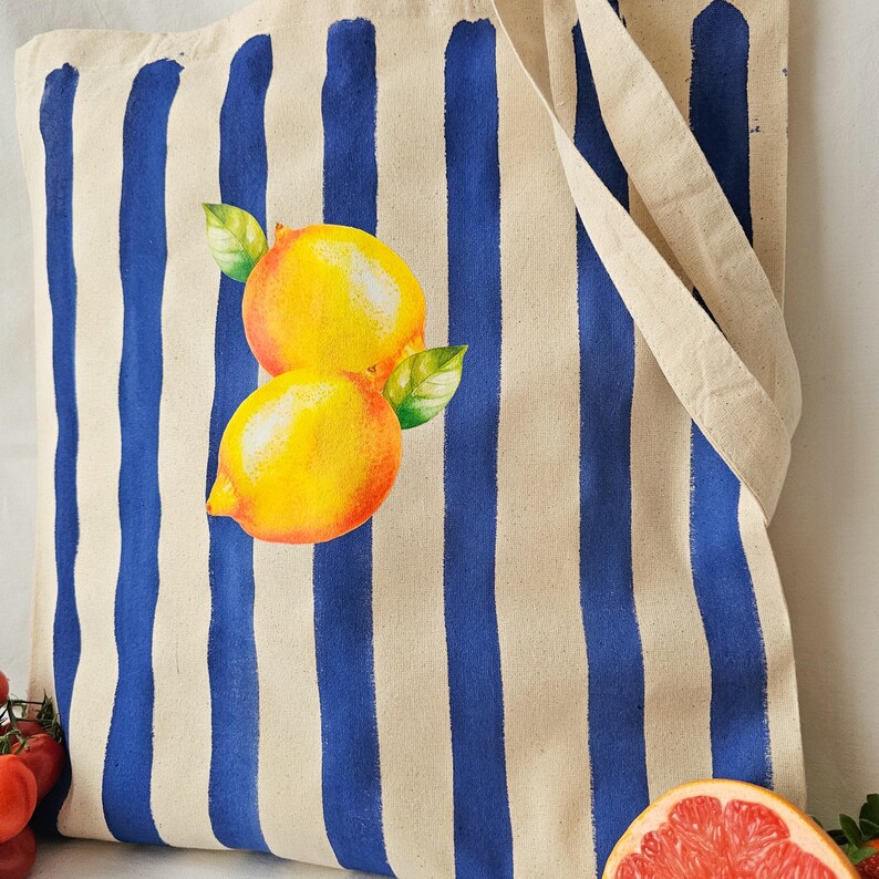 May include: A white canvas tote bag with blue stripes and a painted image of two lemons. The lemons are yellow with green leaves.