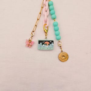 May include: A multi-strand charm accessory featuring a gold-tone metal chain with a pink butterfly charm, a light pink plastic chain with a light blue cassette tape charm, and a strand of light blue beads ending in a gold-tone sunburst coin charm.