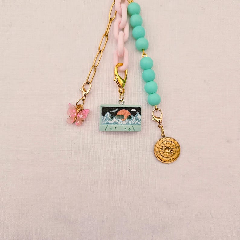 May include: A multi-strand charm accessory featuring a gold-tone metal chain with a pink butterfly charm, a light pink plastic chain with a light blue cassette tape charm, and a strand of light blue beads ending in a gold-tone sunburst coin charm.