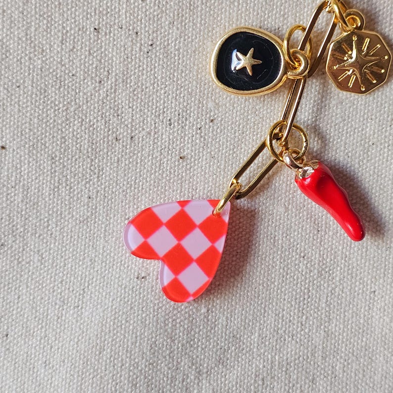 May include: A gold-toned charm necklace with a safety pin clasp. Charms include a red and white checkered heart, a red chili pepper, a black starburst, and a gold compass. The necklace is displayed on a neutral fabric background.