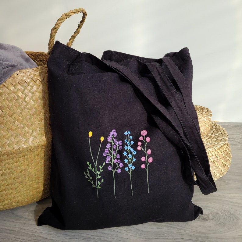May include: Black canvas tote bag with embroidered floral design. The flowers are in shades of purple, blue, pink, and yellow.