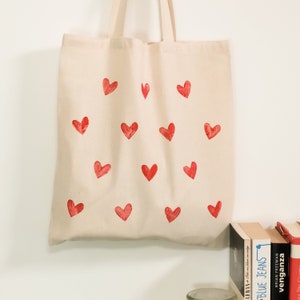 May include: A beige canvas tote bag with a red heart pattern. The bag has a simple design and is perfect for carrying everyday essentials.