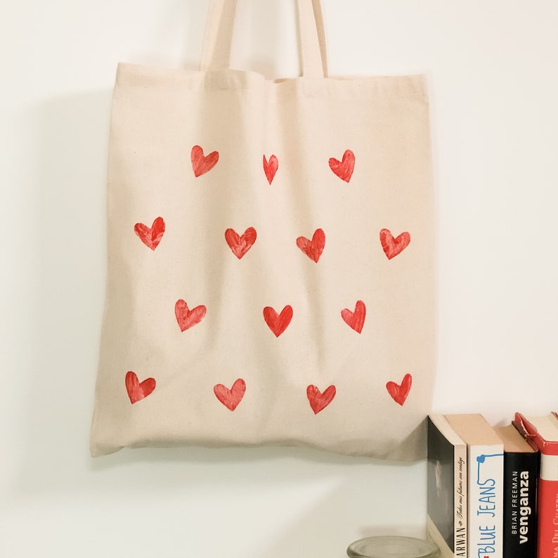 May include: A beige canvas tote bag with a red heart pattern. The bag has a simple design and is perfect for carrying everyday essentials.