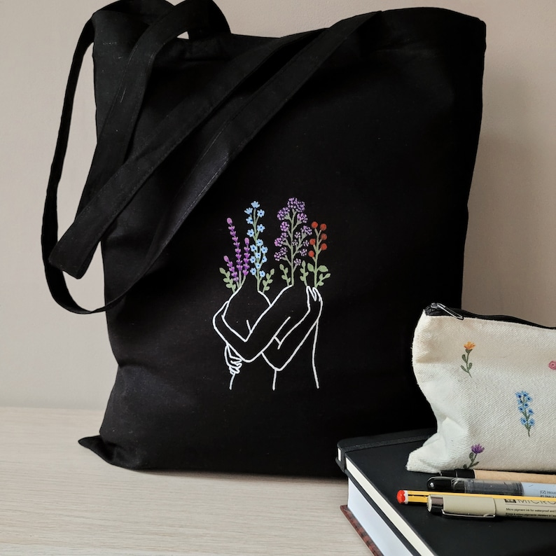May include: Black tote bag with a white line drawing of two figures embracing, with colorful flowers growing from their heads. The bag has a long strap and is made of canvas.
