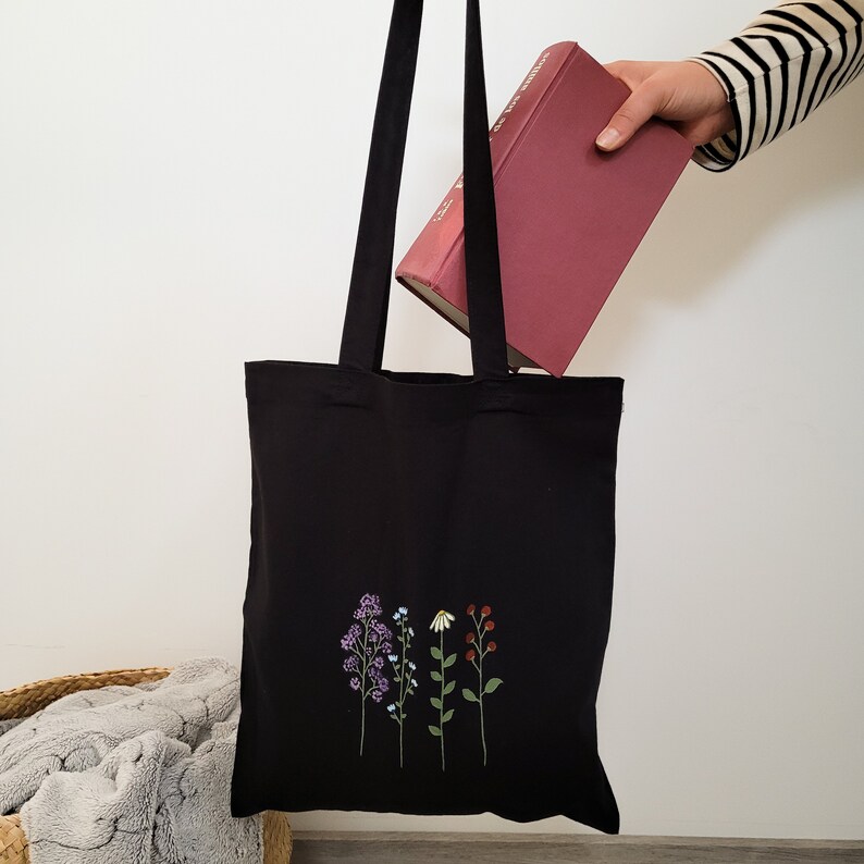 May include: Black tote bag with a floral design embroidered on the front. The flowers are purple, blue, white, and red. The bag is being held by a hand holding a red book.