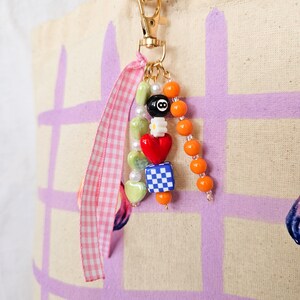 May include: A beaded charm with a gold-colored clasp and a pink and white gingham ribbon. The charm features a variety of beads, including green heart-shaped beads, a red heart, a blue and white checkered cube, and an eight-ball bead.